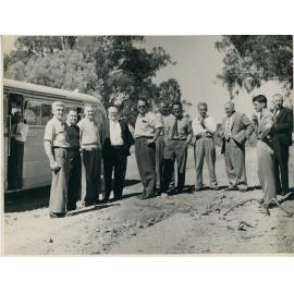 Group of men looking at dirt road with bus in background 